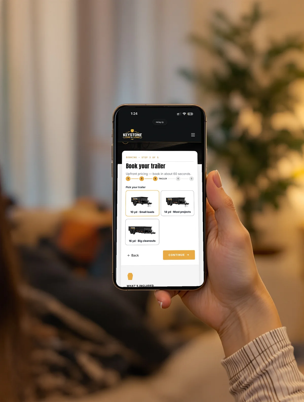 A customer at home on the couch booking on her phone at relay.ly/keystone — Keystone Load & Leave's booking flow at step 3 of 5, picking between 10, 14 and 16 yard trailers, with a Continue button.