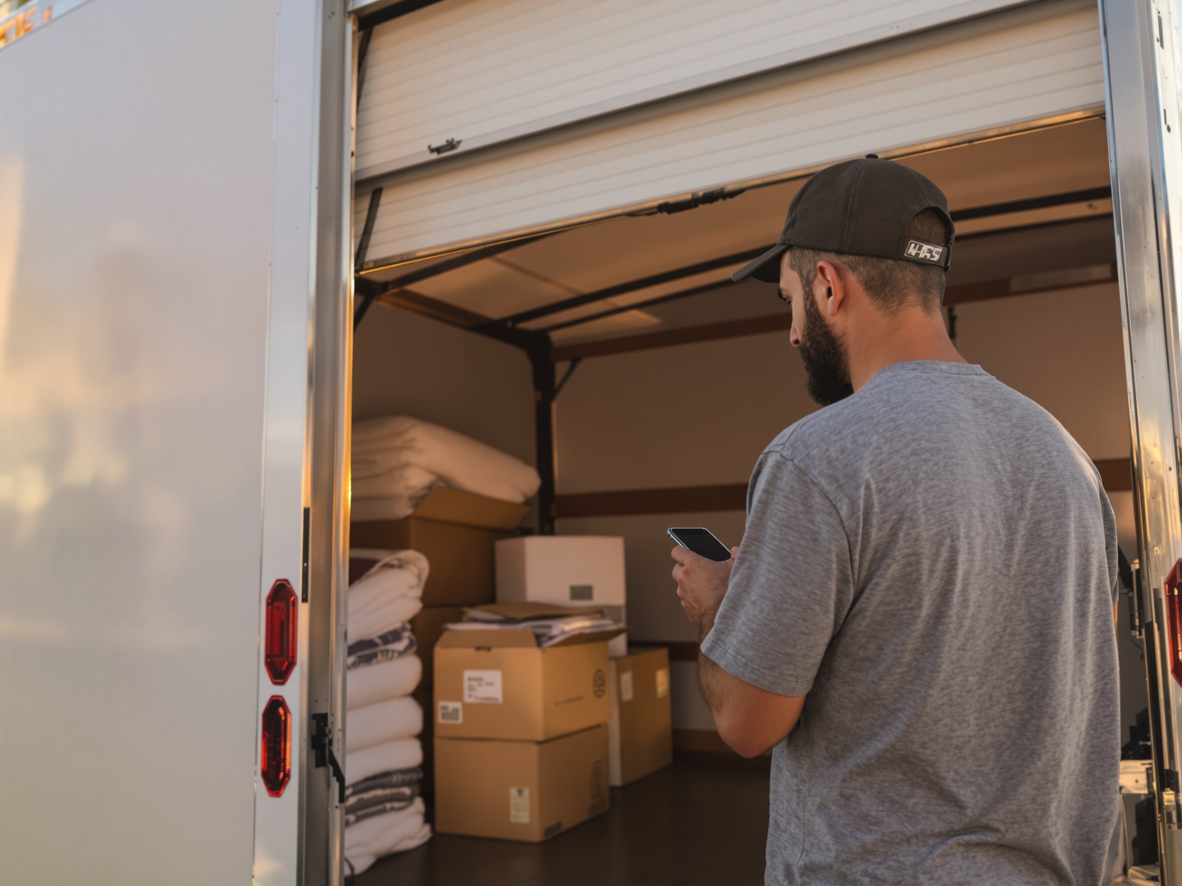 Photo: a mover in a branded work tee closes the roll-up door of a box truck at golden hour, glancing at the phone in his other hand, which shows a Relay new-lead notification. Candid, warm light, shallow depth of field, shot from slightly behind so the truck and phone are both in frame.